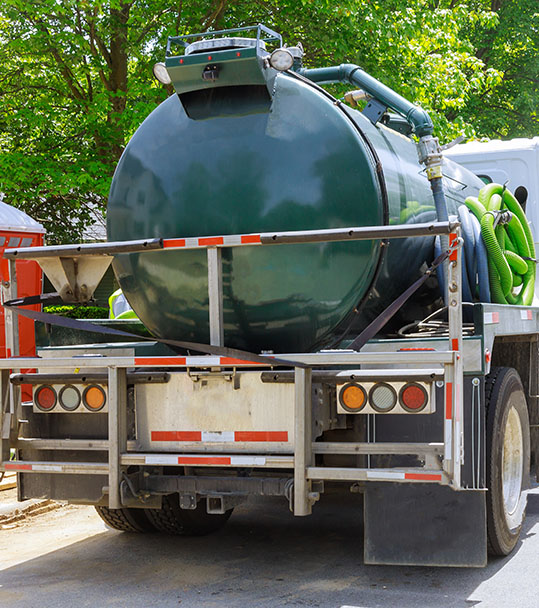 Green vacuum truck with a large cylindrical tank and hoses parked on a tree-lined street. Bright day, clear visibility.