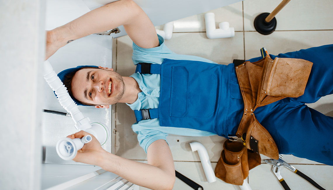 A plumber works beneath a sink, holding tools and examining pipes, with various plumbing parts scattered around the area.