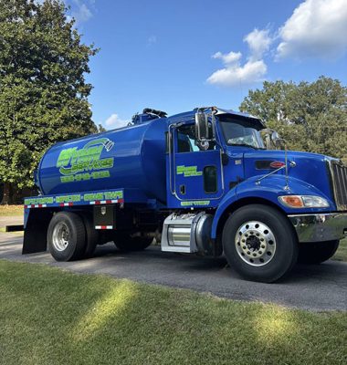 A blue service truck with green lettering, parked on a grassy area with trees in the background under a partly cloudy sky.
