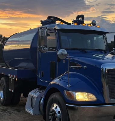 A blue tanker truck is parked on a rural road at sunset, with vibrant clouds and trees in the background.