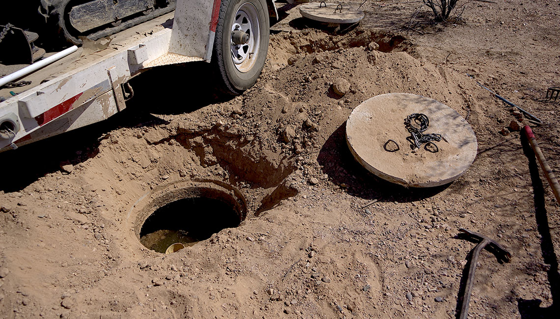 A partially dug hole revealing a circular cover nearby, with tools scattered around on sandy ground and a truck wheel visible.