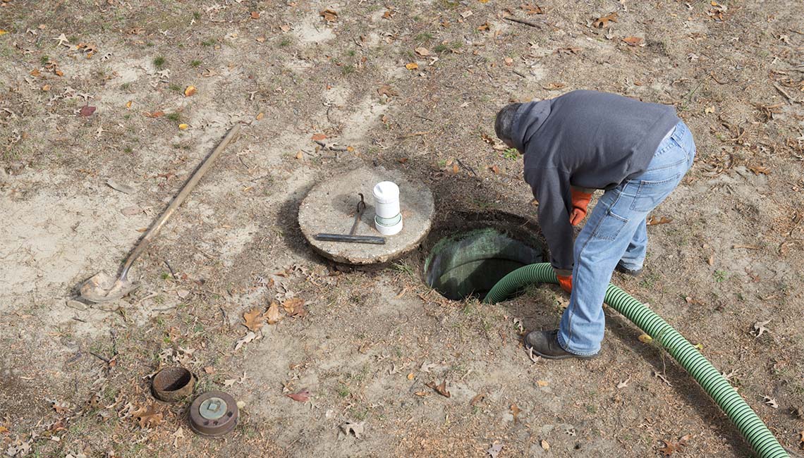 A person in a hoodie leans over an open septic tank, with tools and a green hose nearby, on a dry, leaf-strewn patch of ground.